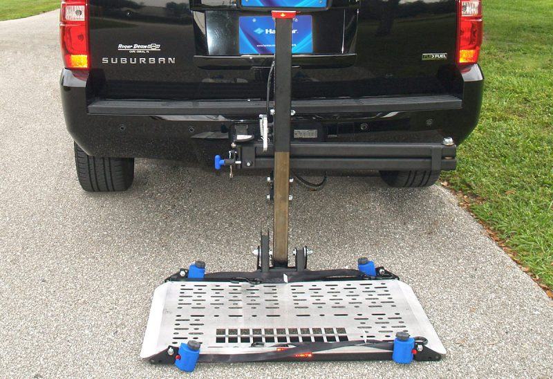 A powered platform lift on a pickup truck's tow hitch, lowered to the driveway.