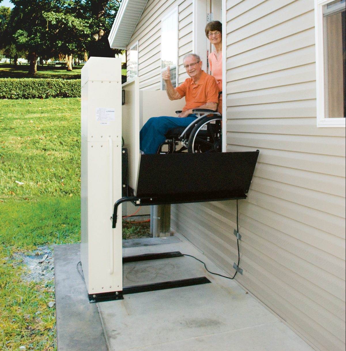 A vertical platform lift mounted beside a house, a man in a manual wheelchair riding the platform up to the door.