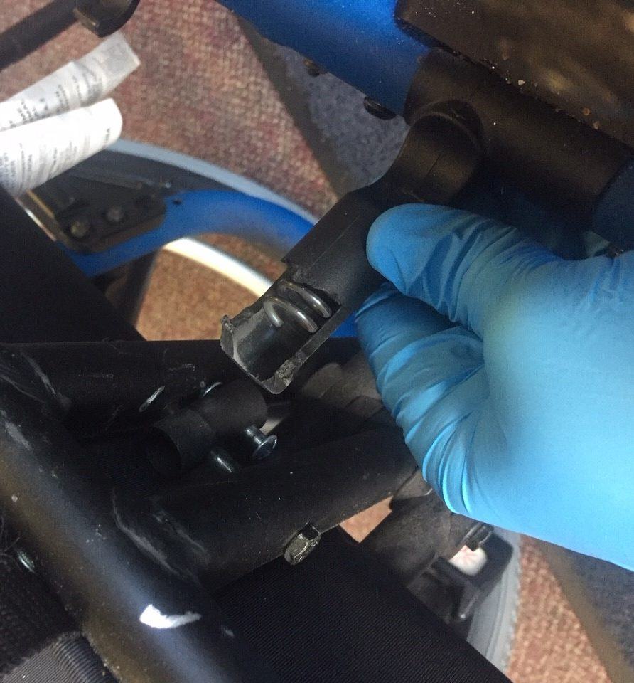 A gloved hand holding a snapped frame attachment point from a manual wheelchair, the broken end and its spring visible, with the chair's blue frame tube behind it.
