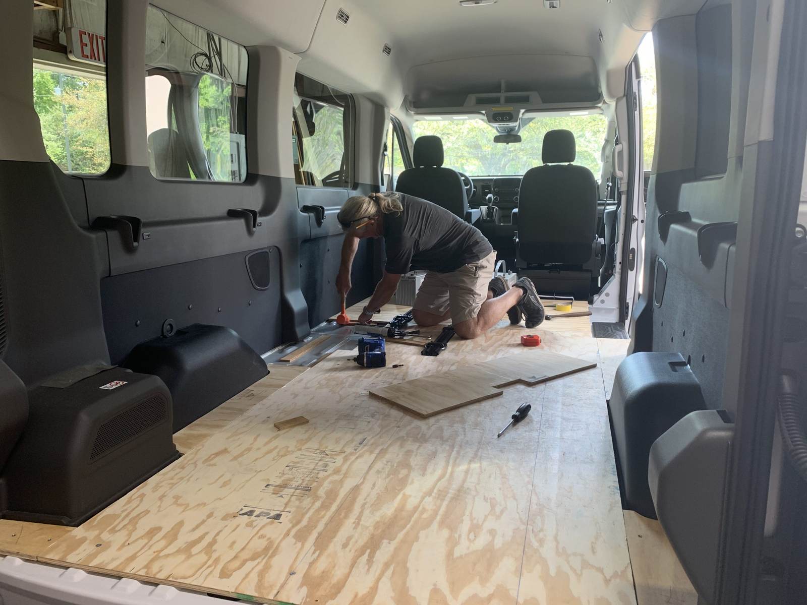 Steve Johnson, the owner of Gator Custom Mobility, kneeling inside a stripped van and fastening the plywood subfloor that a wheelchair conversion is built on. Track rails, a drill and a tape measure lie on the deck around him.